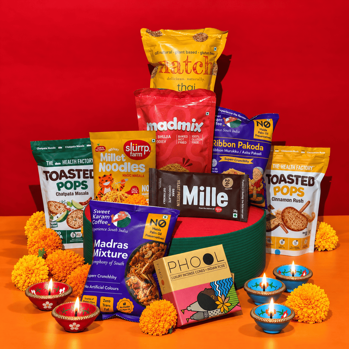 A festive snack and treat gift arrangement featuring multiple packaged food items including Match Thai, Millar Noodles, Slurr Mixture, Mille chocolate, Ribbon Pakoda, and Toasted Pops, styled with marigold flowers and lit diya candles against a red and orange backdrop.