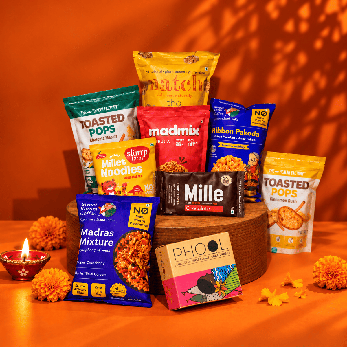 A styled snack and coffee gift assortment arranged on a wooden pedestal against a warm orange background. Visible products include Matcha Matcha tea, toasted popcorn, miller noodles, slurpee, Mille chocolate, Madras Mixture, PHOOL tea, and Ribbon Pakoda, with marigold flowers and a small lit diya around them.